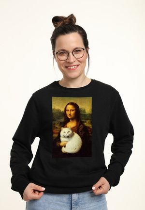 Lächelnde Frau mit Brille und Dutt trägt einen schwarzen Pullover mit der Mona Lisa, die eine weiße Katze hält.