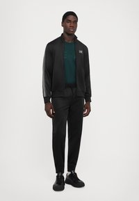 Man wearing black zip-up track jacket with stripes on sleeves, black track pants, black sneakers, and a dark green logo t-shirt, standing on white background.