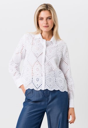 Witte geborduurde blouse met een gegolfde zoom, voorzien van ingewikkelde gatenpatronen, lange mouwen en knoopsluiting. Gecombineerd met blauwe denim broek.
