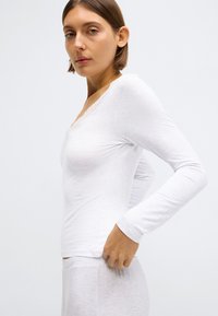 Biely top s dlhým rukávom s výstrihom do V a čipkovanou výzdobou, priliehavý strih a mäkký, textúrovaný materiál. Modelka upravuje lem.