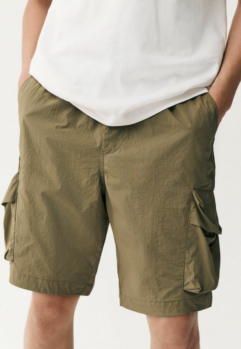 Homme portant un short cargo vert olive longueur genoux avec poches latérales et un t-shirt blanc, mains dans les poches, debout devant un fond uni.