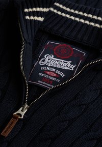 Navy blue cable-knit sweater with a zipper, leather zipper pull, white stripes on collar, and a Superdry brand label inside.