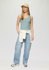 Hellblaues, geripptes Tanktop mit Spitze, kombiniert mit lockeren, hellen Denim-Cargohosen und einer beigen Mütze, mit einem cremefarbenen Pullover um die Taille gebunden.