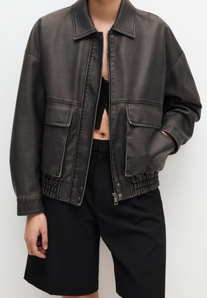 Person iført en mørkebrun læderjakke med lommer foran, delvist åbnet lynlås, over sorte cropped bukser, stående mod en ensfarvet baggrund.