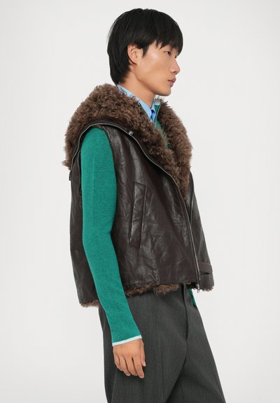Young person wearing teal sweater, blue shirt, gray pants, and dark brown leather vest with large fluffy collar, standing in profile.