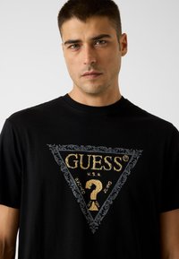 Zwart T-shirt met korte mouwen met een driedelig gouden logo met sierlijke details, merktekst en een vraagtekenafbeelding.