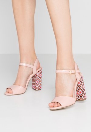 Różowe lakierowane sandały na obcasie z otwartym palcem, paskiem wokół kostki oraz blokowym obcasem w geometryczny wzór w odcieniach różu, czerwieni i czerni.