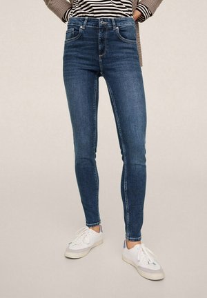 Person iført mørkeblå skinny jeans, hvide sneakers, stribet skjorte og ternet blazer med hænderne i lommerne mod en ensfarvet baggrund.