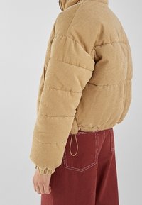 Chaqueta acolchada de pana en color beige con puños y dobladillo elásticos. Presenta un cuello alto y pantalones rojos contrastantes con costuras blancas.