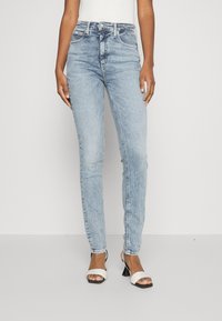 Jeans de tiro alto en azul claro con un ajuste slim, hechos de mezclilla. Presentan un diseño clásico de cinco bolsillos y un desvanecimiento sutil. Se combinan con sandalias de tacón blancas.