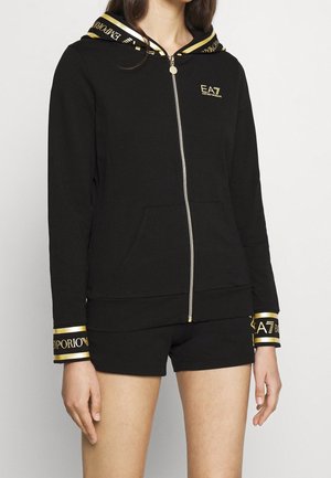 Sweat à capuche zippé noir avec poignets et capuche bordés de doré, logo EA7 Emporio Armani doré sur la poitrine, porté avec un short noir.