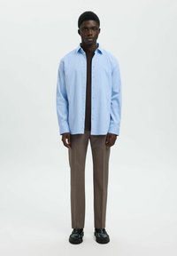 Jasnoniebieska koszula zapinana na guziki, ciemny golf pod spodem, brązowe dopasowane spodnie, czarne buty; gładki materiał, dopasowany krój i naturalna sylwetka.