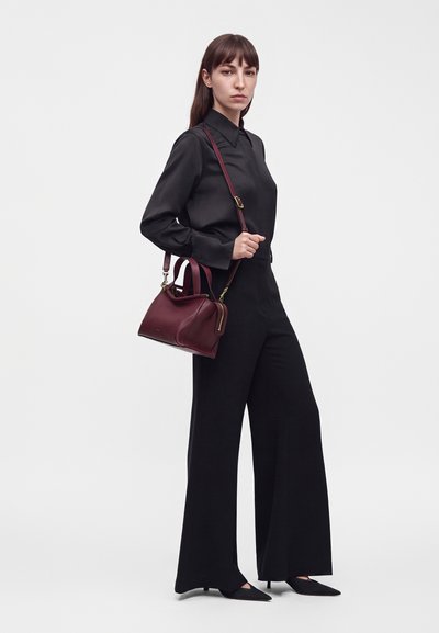 Femme portant un chemisier noir à manches longues et un pantalon large, tenant un sac à main en cuir rouge foncé avec une bandoulière sur un fond blanc uni.