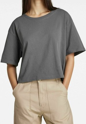 Person wearing a loose dark gray cropped t-shirt with short sleeves and light beige pants with hands in pockets.