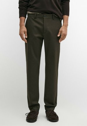 Uomo che indossa pantaloni marrone scuro a gamba dritta, cintura nera, scarpe marrone scuro con lacci e camicia scura a maniche lunghe.