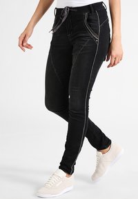 Pantalones jogger de denim negro con cintura ajustable, costuras en beige contrastantes y bolsillos laterales. Llevados con zapatillas de color claro.
