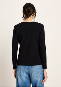 Frau mit dunklem, geflochtenem Haar, die ein einfaches schwarzes Langarmshirt und hellblaue Jeans trägt und mit dem Rücken zur Kamera steht.