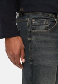 Jean en denim foncé avec un motif légèrement délavé, présentant des coutures contrastées et une poche avant. Main reposant sur le côté.