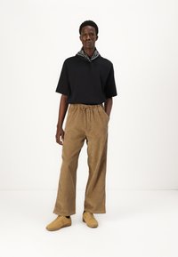 Sort kortærmet t-shirt, brune kordfløjsbukser med vidde og beige sneakers. Sort-hvid mønstret tørklæde knyttet om halsen.