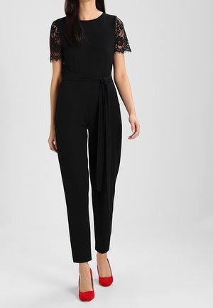 Sort jumpsuit med korte ærmer i blonder, tætsiddende silhuet og bælte i taljen. Kombineret med røde højhælede sko. Glat tekstur.