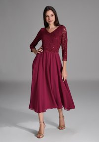 Robe bordeaux avec un haut en dentelle, décolleté en V et manches 3/4. Jupe en chiffon fluide avec un ourlet au niveau des genoux, complétée par des sandales dorées.