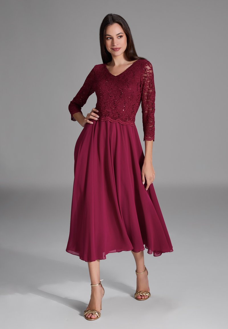 Robe bordeaux avec un haut en dentelle, décolleté en V et manches 3/4. Jupe en chiffon fluide avec un ourlet au niveau des genoux, complétée par des sandales dorées.