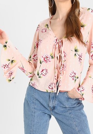 Hellrosa Bluse aus blümchenmusterndem Stoff mit Glockenärmel und gebundenem Halsdetail, kombiniert mit hochtaillierten blauen Denim-Jeans.