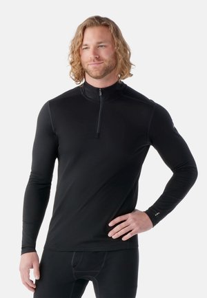 Hombre con cabello rizado hasta los hombros, vestido con una prenda negra de manga larga con cremallera y pantalones negros, de pie con una mano en la cadera.