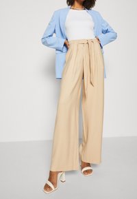 Lichtblauw colbert over een wit topje, gecombineerd met beige, hooggetailleerde, geplooide broek en witte sandalen met hak. Glad stoftextuur.