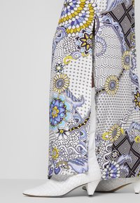 Pantalon long à motifs mélangeant des designs géométriques et floraux en blanc, bleu, jaune et noir, associé à des bottines blanches texturées.