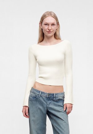 Jonge vrouw met lang blond haar, gekleed in een aangesloten witte geribbelde top met lange mouwen en losse blauwe spijkerbroek tegen een effen achtergrond.