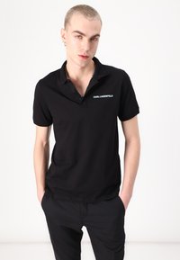 Polo nero in cotone, con collo classico, maniche corte e logo sul lato sinistro del petto. Indossato con pantaloni neri.