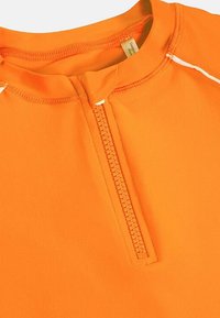 Top deportivo naranja con una cremallera corta, costuras planas y un cuello alto. Está hecho de una tela suave y elástica con acentos blancos.