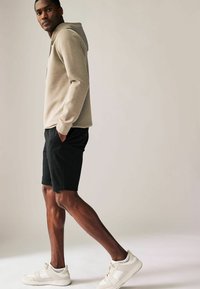 Hombre de pie con las manos en los bolsillos, vestido con sudadera con capucha beige, pantalones cortos negros y zapatillas blancas sobre un fondo liso.