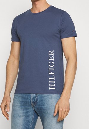 Mężczyzna w dopasowanej granatowej koszulce z krótkim rękawem, z białym pionowym napisem "HILFIGER" po lewej stronie, w połączeniu z jasnoniebieskimi dżinsami.