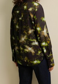 Chemise boutonnée en tissu foncé avec un motif floral en verts et blancs. Comprend un pli dans le dos et des manches longues avec poignets.