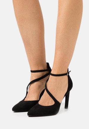Chaussures à talons hauts en daim noir avec des bouts pointus et des lanières fines autour des chevilles croisant les pieds sur les jambes d'une personne.