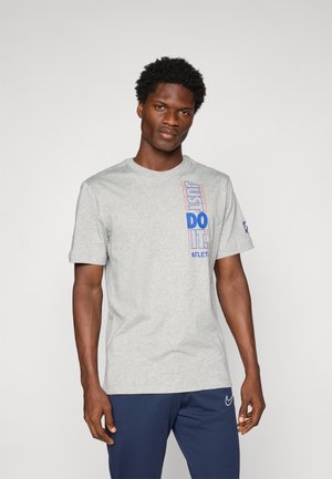 Man in een lichtgrijze T-shirt met blauwe en rode tekst "Just Do It" en marineblauwe broek tegen een effen lichte achtergrond.