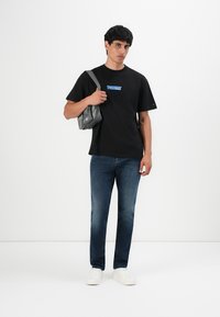 Jonge man in een zwart "Tommy" logo t-shirt, blauwe jeans, witte sneakers, met een grijze schoudertas tegen een witte achtergrond.