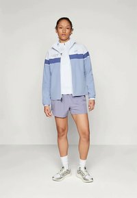 Jaqueta atlética azul clara com listras brancas e azul-marinho, combinada com calções cinzentos. O modelo usa meias brancas e ténis cinzentos com acabamentos texturizados.