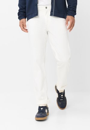 Persoon draagt witte broek, marineblauwe sneakers met rubberen zolen en een marineblauwe jas, lopend op een effen witte achtergrond.