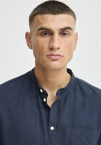 Camisa de botón azul oscuro con cuello mandarín, bolsillo en el pecho y botones blancos. Confeccionada con una tela texturizada y un diseño casual.