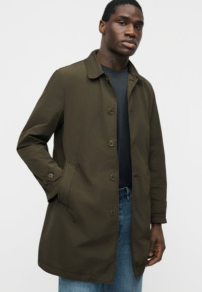 Άνδρας ντυμένος με παλτό ελιάς με κουμπιά και πλαϊνές τσέπες πάνω από σκούρο πουκάμισο και μπλε τζιν, στεκόμενος μπροστά σε λευκό φόντο.