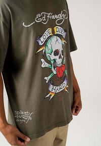 Olive green t-shirt featuring a large graphic of a skull with crossbones, dripping heart, and colorful banner stating "LOVE DIES HARD."