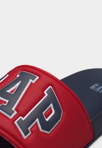 Gros plan sur une sandale à glissière rouge de la marque GAP avec de grosses lettres grises et blanches sur la sangle, posée sur une semelle sombre.