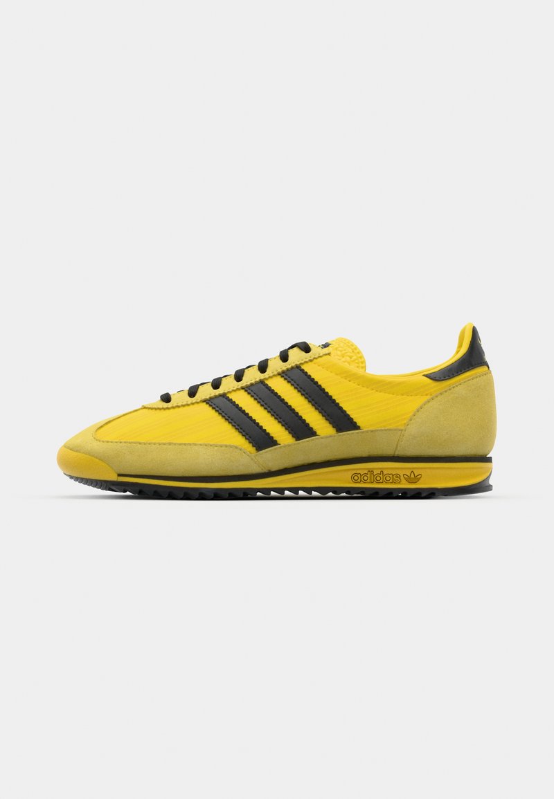 Zapatillas deportivas amarillas con franjas negras, de materiales de ante y malla, diseño de perfil bajo, suela de goma y detalle del logo en el talón.