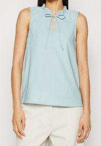 Top sin mangas de color azul claro con cuello con volantes y un escote con forma de llave, que presenta un cierre de lazo. Tejido suave y ajuste holgado.