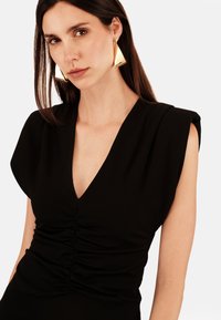 Černé šaty s výstřihem do V a nabíran�ými detaily v pase. Modelka nosí velké trojúhelníkové zlaté náušnice. Hladká tkanina, strukturované ramena.