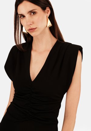 Czarna sukienka z dekoltem w kształcie litery V i marszczeniami w talii. Modelka nosi duże, trójkątne złote kolczyki. Gładki materiał, usztywnione ramiona.