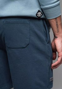 Granatowe szorty wykonane z miękkiego materiału, z tylną kieszenią z zaokrąglonym klapkiem oraz małą metką z logo blisko pasa.